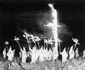 Picture of a burning cross in a field surrounded by white clad and hooded Ku Klux Klan members