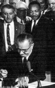 President Lyndon B. Johnson signing a document with Martin Luther King Jr. looking on